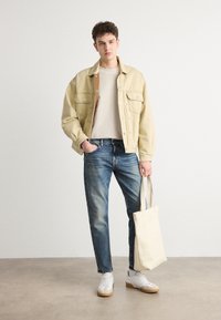Jachetă din denim bej, tricou crem, blugi albaștri, purtând o geantă de umăr neutră. Adidași albi cu talpă bej. Îmbrăcăminte simplă și lejeră.