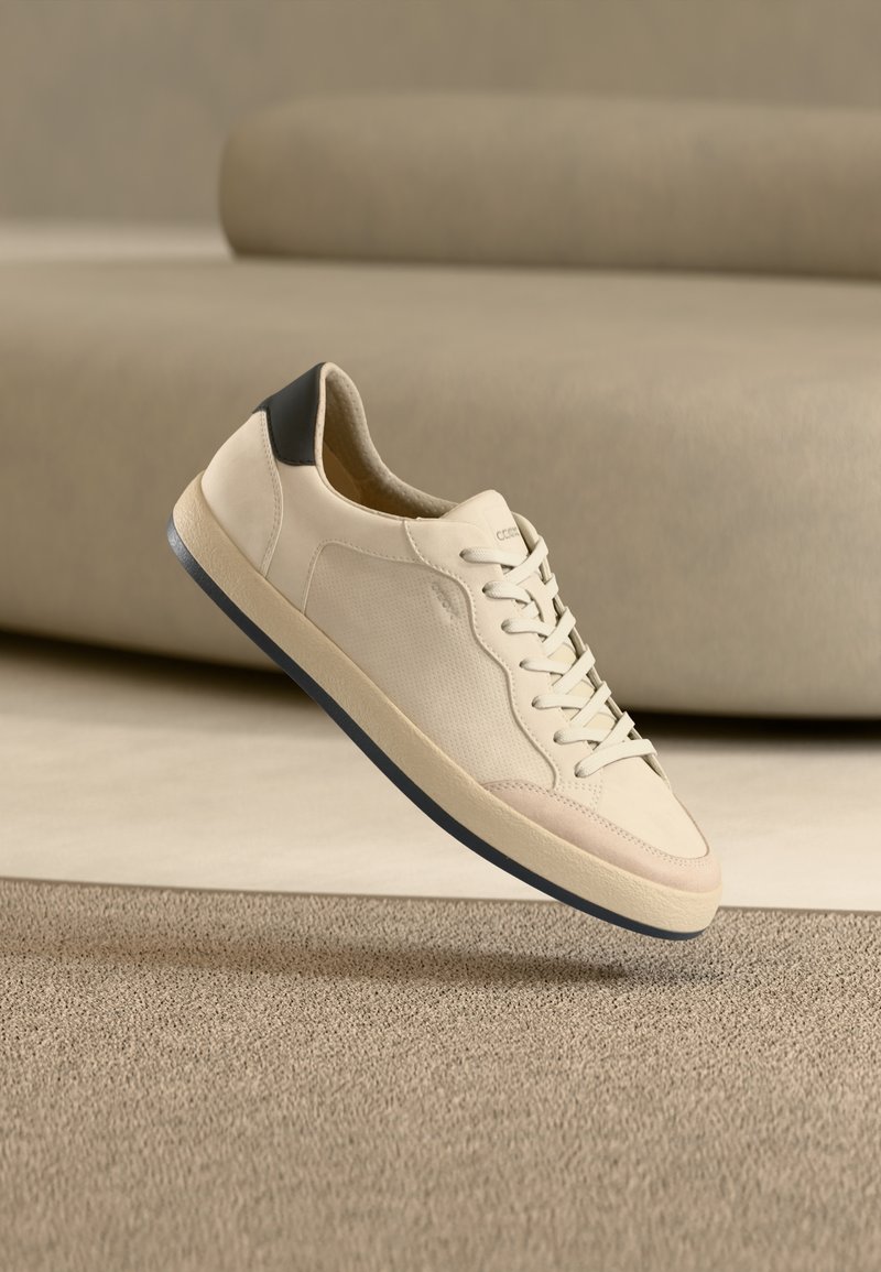 Μπεζ δερμάτινο αθλητικό παπούτσι με μαύρη λεπτομέρεια στη φτέρνα, στρογγυλή μύτη και επίπεδη ελαστική σόλα. Διαθέτει ραφές στο ίδιο χρώμα και σχεδιασμό με κορδόνια.