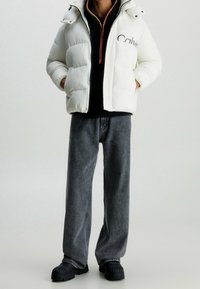 Doudoune blanche avec détail logo, polaire noire zippée en dessous, jean gris à jambes larges, et bottes noires à semelle épaisse.