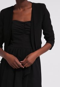 Černý zkrácený blejzr s t�říčtvrtečními rukávy, strukturovaným designem a bez knoflíků, vrstvený přes černé šaty s nabíraným korzetem.