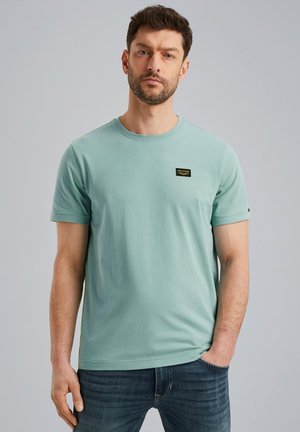 Minzgrünes Baumwoll-T-Shirt mit Rundhalsausschnitt, kurzen Ärmeln und einem kleinen schwarzen Etikett über der linken Brust. Kombiniert mit blauen Jeans.