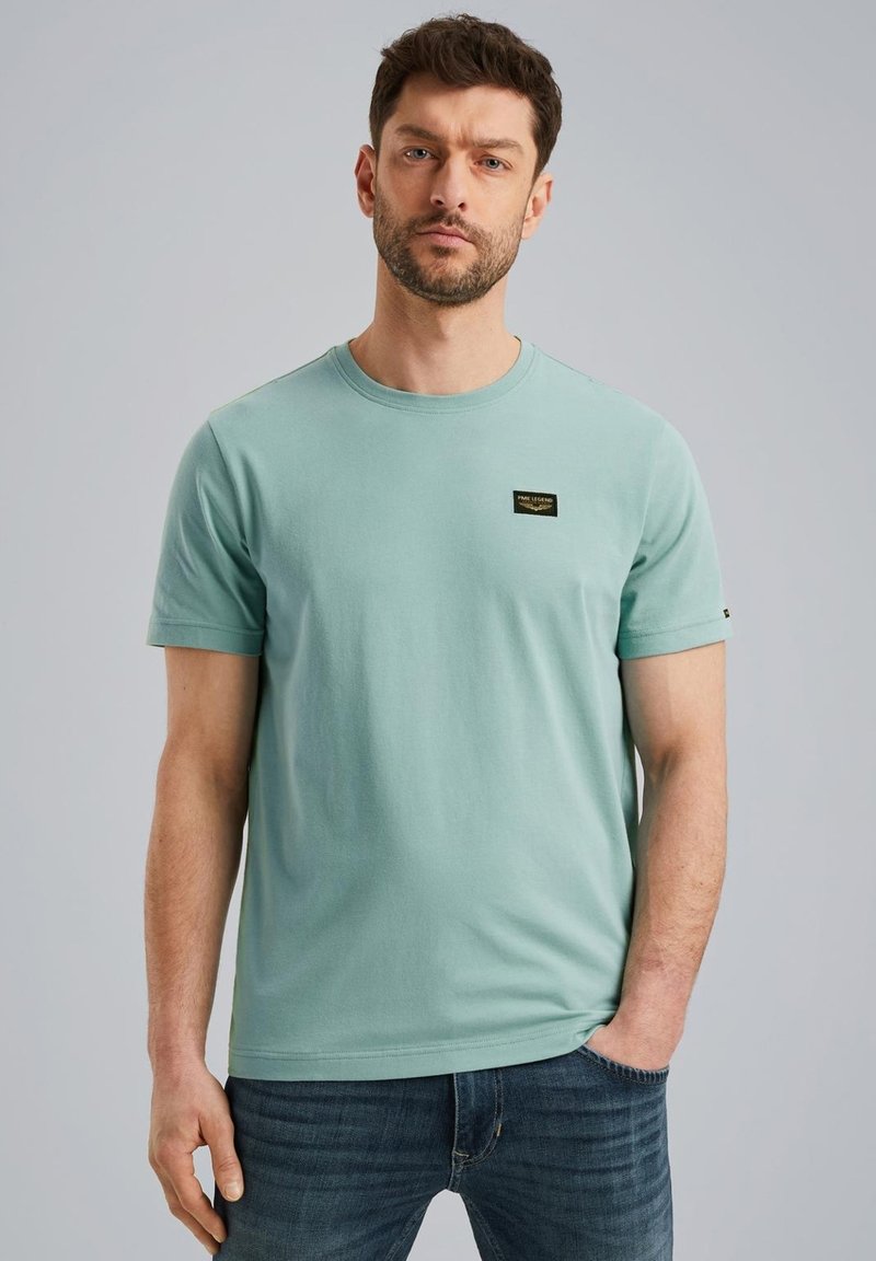 Camisa de algodón de color verde menta con cuello redondo, mangas cortas y una pequeña etiqueta negra sobre el lado izquierdo del pecho. Combinada con jeans azules.
