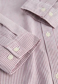 Gros plan d'une chemise à manches longues pliée avec des rayures verticales bordeaux et blanches et des boutons blancs sur le poignet et la patte de boutonnage avant.