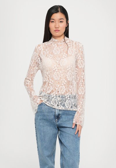 Kanten top met lange mouwen in gebroken wit met bloemenpatroon, hoge hals, transparant ontwerp en uitlopende mouwen, gecombineerd met blauwe denim jeans.
