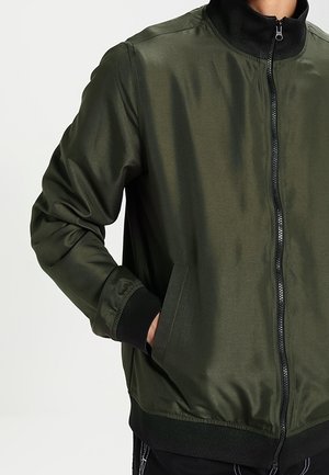 Mann trägt eine olivgrüne Bomberjacke mit schwarzem Kragen, Manschetten, Bund und Frontreißverschluss, Hand in der linken Tasche.
