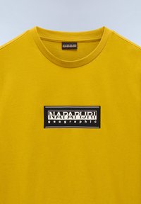 Gul bomulds T-shirt med rund hals, korte ærmer og et sort rektangulært logo-patch med hvid tekst centreret på forsiden.