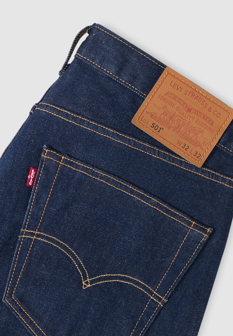Dark blue denim jeans with a leather patch on the waistband. Features contrast yellow stitching and a red Levi's tab on the back pocket.