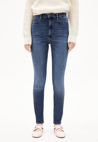 Vaqueros ajustados de mezclilla azul oscuro, de cintura alta, con bolsillos delanteros y una textura suave. Combinados con zapatos planos de color rosa claro.