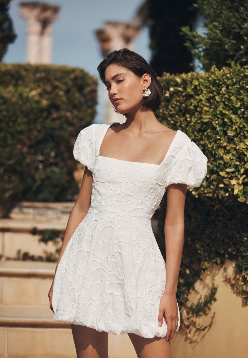 Weißes Kleid mit Puffärmeln und quadratischem Ausschnitt, verziert mit strukturierten floralen Stickereien. Kurze, ausgestellte Form, vor grünem Hintergrund.