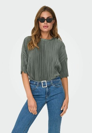 Top verde con pliegues y mangas cortas, combinado con vaqueros de tiro alto y un cinturón blanco. El modelo lleva gafas de sol negras y está posando.