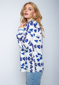 Biała bluzka w niebieskie wzory serc, o luźnym kroju, z niebieskimi paskami na mankietach. Gładki materiał z lekkim połyskiem.