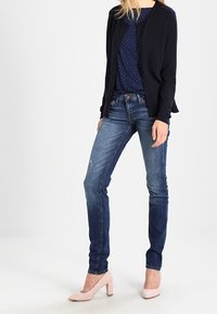 Cárdigan azul marino sobre una blusa azul marino con lunares, combinado con vaqueros azules y zapatos con tacón cuadrado de color rosa claro. Tela suave y diseño ajustado.