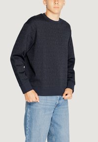 Felpa blu navy con un motivo a logo testurizzato su tutta la superficie, polsini e scollatura a coste, abbinata a jeans azzurri chiari.