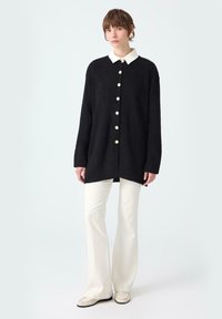 Woman wearing black oversized buttoned cardigan with white collar, cream flared pants, and cream loafers standing on plain background.