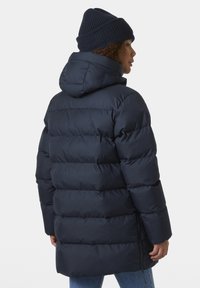 Chaqueta tipo puffer azul marino con capucha, con secciones acolchadas en horizontal y cremalleras laterales. Combinada con un gorro de punto azul marino.