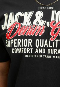 Schwarzes Baumwoll-T-Shirt mit fettgedrucktem weißen und roten Text, der "JACK & JOE Denim G" und "ÜBERLEGENE QUALITÄT, KOMFORT UND HALTBARKEIT" liest.