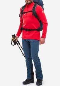 Homme portant une veste rouge et un pantalon de randonnée bleu, portant un sac à dos bleu et tenant une paire de bâtons de trekking, se tenant contre un fond blanc.