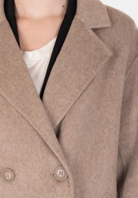 Cappotto doppiopetto beige in misto lana con rever a lancia e due bottoni, caratterizzato da una texture liscia e una vestibilità strutturata, parzialmente visibile.