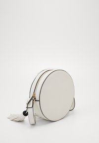 Kulatá bílá kabelka přes rameno s texturovaným povrchem, černým okrajem, zlatým zipem a třásňovým zdobením. Kompaktní a elegantní design.