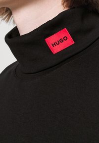 Schwarzes Rollkragen-Shirt aus weichem Stoff, mit einem roten Quadratlabel mit dem "HUGO"-Logo am Kragen. Glatte Textur und schlichtes Design.