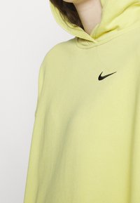Felpa gialla con logo Nike nero sul petto, caratterizzata da una texture morbida e un cappuccio. La vista ravvicinata mostra i dettagli del collo e delle spalle.