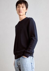 Marineblauer, strukturierter Strickpullover mit V-Ausschnitt und langen Ärmeln, kombiniert mit hellblauen Jeans. Der Stoff wirkt weich und lässig.