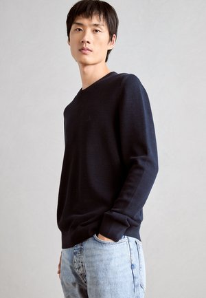 Marineblauer, strukturierter Strickpullover mit V-Ausschnitt und langen Ärmeln, kombiniert mit hellblauen Jeans. Der Stoff wirkt weich und lässig.