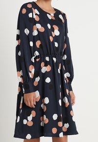 Granatowa sukienka z długimi rękawami, zwężona w talii, z wielokolorowym, okrągłym wzorem w kropki w kolorze brzoskwiniowym, czarnym i białym.