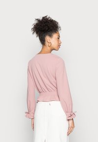 Pink long-sleeve top with a cinched waist, gathered sleeves with ties, and a rounded neckline, paired with a white skirt.