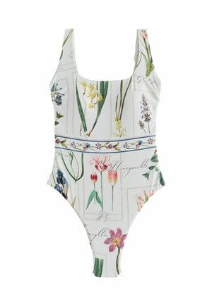 Lyshåret kvinde i en blomsterprintet badedragt står ved store sten med hav og himmel i baggrunden og kigger mod venstre.