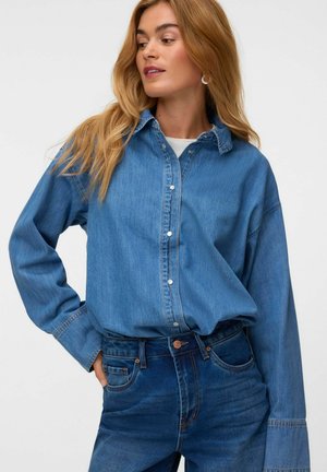 Frau trägt ein mittelblaues Jeanshemd mit Knöpfen, das in hoch taillierte, dunklere blaue Jeans gesteckt ist, und schaut nach links.