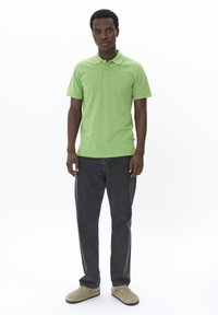 Ljusgrön polo med treknappsslutning, mörk denimjeans och beige slip-on skor. Enfärgad bakgrund, modell stående upprätt.