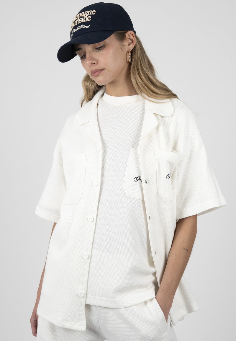 Femme portant une chemise blanche texturée à manches courtes boutonnée sur un haut et un pantalon assortis, avec une casquette de baseball bleu marine, les mains dans les poches, regardant vers le bas.