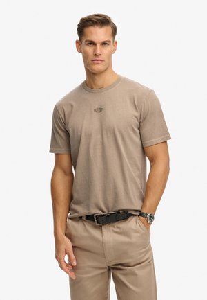 Jasnobrązowa bawełniana koszulka z krótkimi rękawami i okrągłym dekoltem. Posiada subtelny nadruk logo na klatce piersiowej. Noszona z jasnobeżowymi spodniami.