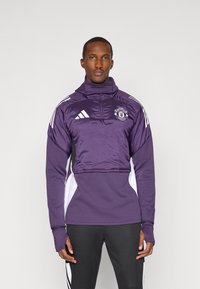 Lilla træningsjakke med høj krave, lynlåslukning foran, hvide sidestykker og Manchester United-logo; har tekstureret stof og lange ærmer.