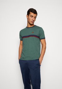 Camiseta de algodón verde con una franja horizontal en azul marino y burdeos. Cuenta con un cuello de canalé y un pequeño logo en el pecho.