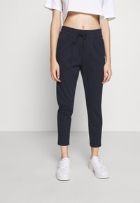 Marineblå smale bukser med snor i livet, laget av et glatt stoff. Kombinert med hvite sneakers for et avslappet utseende.