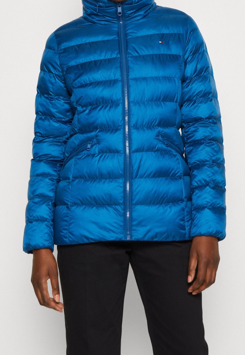 Personne portant une veste matelassée bleu vif avec une fermeture éclair frontale et des poches zippées, associée à un pantalon noir, sur un fond uni.