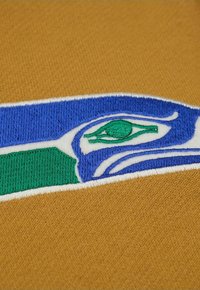 Bestickter Stoff mit braunem Hintergrund; zeigt ein blau-grünes Design mit einer Augenform und stilisierten Wellenmustern in Weiß.