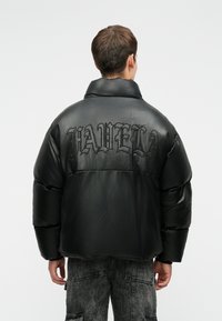 Fekete puffjacket fényes felülettel, amely oversized szabással, magasGallérral és hímezett szöveggel rendelkezik a hátsó részén.