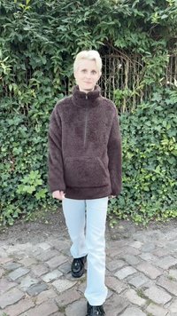Pulóver de forro marrón con cuello alto y cierre de cremallera, combinado con pantalones azul claro y zapatos negros, en un entorno de frondosa vegetación verde.