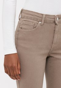 Jeans de mezclilla beige con una cintura de altura media, que cuentan con un cierre de botón, presillas para el cinturón y bolsillos frontales. Textura suave con sutiles detalles de costura.