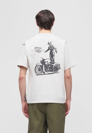 Persoon draagt een wit T-shirt met een zwart print van een man die op een vintage motorfiets staat en de tekst "Dickies" op de achterkant.