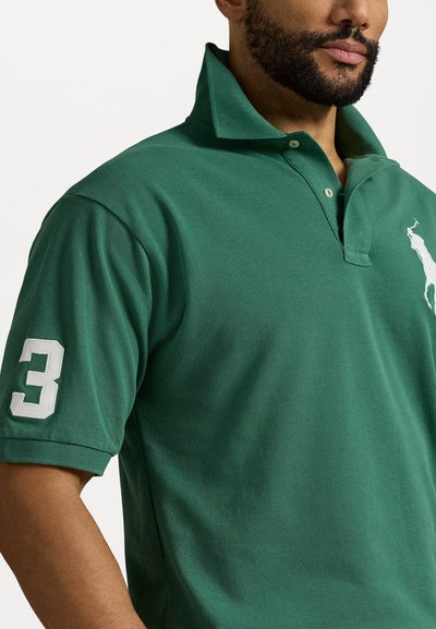 Žalias polo marškinis, pagamintas iš tekstūruoto audinio, su trimis mygtukais nesugadintu apykakle, balta siuvinėta logotipu ir dideliu baltu numeriu 3 ant rankovės.