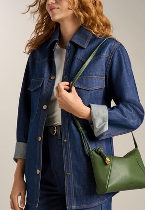 Femme portant une veste en jean foncé et un jean avec un haut blanc, tenant un sac à bandoulière en cuir vert avec une sangle nouée.