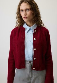 Czerwony sweter z dzianiny z teksturowanym wzorem w romby, ze ściągaczami i srebrnymi guzikami, noszony na jasnoniebieskiej koszuli z kołnierzykiem.