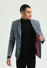 Grau blazer mit strukturiertem Design, ausgestattet mit schwarzen Knöpfen und einem pinken Innenfutter, getragen über einem schwarzen Rollkragenpullover.