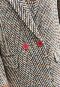Sako na míru v herringbone vzoru. Texturovaná tkanina v šedé barvě s multibarevnými flíčky. Obsahuje dvojité červené knoflíky a boční kapsy.