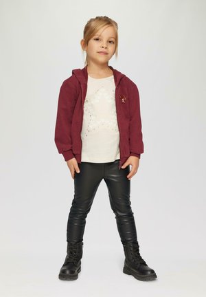 Bordeaux rode zip-up hoodie, crèmekleurig t-shirt met zilveren hartpatroon, zwarte leren legging en zwarte veterschoenen.