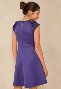 Lila Kleid mit Spitzenakzenten an den Schulterpartien, floralen Details, figurbetontem Oberteil, ausgestelltem Rock und einer Schlüsselaugenöffnung im Rücken mit einem einzigen Knopfverschluss.